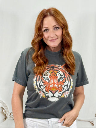 A woman with long wavy red hair wearing a dark gray graphic t-shirt featuring a large, colorful tiger face print on the front, paired with white pants, set against a light gray studio background.