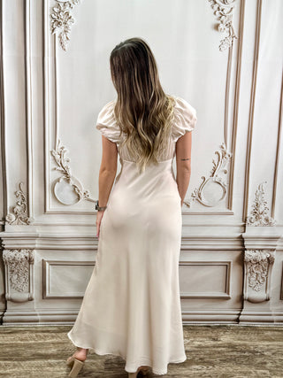 A woman viewed from behind wearing a cream-colored, floor-length slip dress with short puff sleeves, standing in front of an ornate, light-colored paneled wall.
