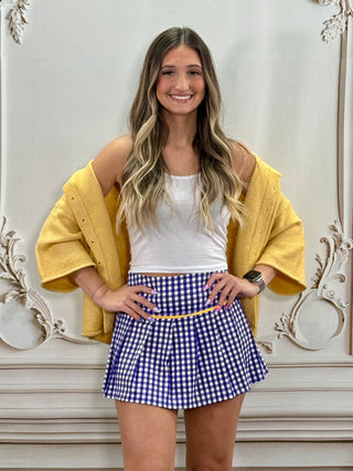 A woman modeling a purple and white gingham pleated mini skirt with a yellow waistband, styled with a white tank top, a yellow knit cardigan, and a watch, standing against an ornate white wall.