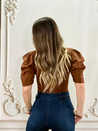 A woman viewed from behind wearing a brown puff-sleeve top and blue jeans, standing against an ornate white wall.