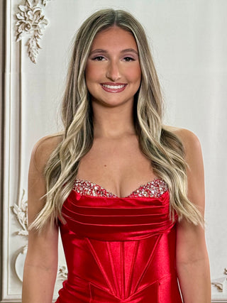 A smiling woman with long blonde-highlighted hair wearing a bright red strapless satin dress with a jeweled embellished sweetheart neckline, posed against an ornate white wall.