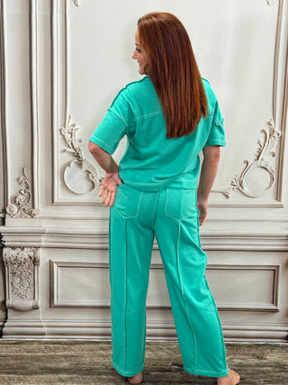 A woman with long red hair is shown from the back wearing a matching turquoise green scrub set, consisting of a short-sleeve top and wide-leg pants, standing in front of an ornate white wall.