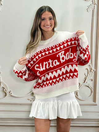 A woman wearing a red and white knit sweater with 'Santa Baby' written across the chest, paired with a white ruffled skirt, standing in front of an ornate white wall.