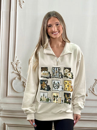 A woman wearing a cream-colored quarter-zip sweatshirt with a graphic print of the word 'LOVE' in Louisiana state silhouettes, posed against a white classical wall.
