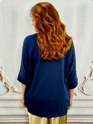 A woman with long wavy red hair is shown from the back, wearing a loose navy blue top with three-quarter sleeves and light gold pants, standing against an ornate white wall.
