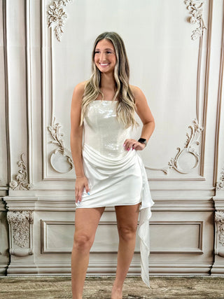 A woman modeling a strapless white sequined mini dress with a draped side detail, standing in front of an ornate beige wall.