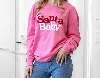Person wearing a pink sweatshirt with 'Santa Baby' text, standing against a white wall.