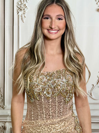 A smiling woman with long blonde-highlighted hair wears a strapless embellished gown in warm beige and gold tones, standing indoors against a decorative cream wall.