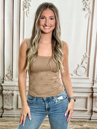 A smiling young woman with long blonde-highlighted hair stands indoors wearing a fitted sleeveless tan top with a subtle tonal floral or textured pattern and blue jeans. The background is an ornate cream-colored wall with decorative molding.
