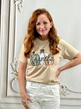 A woman with long red hair wearing a beige graphic t-shirt that reads 'Thankful' in script over three patterned pumpkins, paired with white pants, standing in front of a white decorative wall.