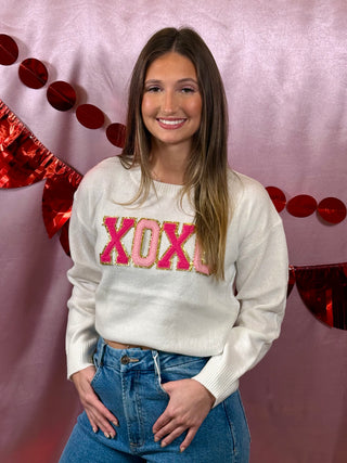 Woman wearing a white sweater with 'XOXO' in pink and red sequins against a pink background with heart decorations.