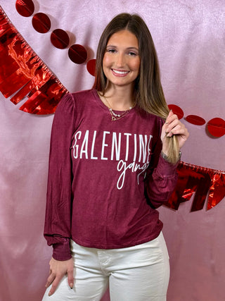 Woman wearing a maroon sweatshirt with 'GALentine gang' text, standing against a pink background with red heart decorations.