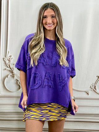 A woman modeling an oversized purple t-shirt with 3D lettering that reads 'Tired of being normal' and a yellow and purple wavy patterned mini skirt, standing against a white decorative wall.