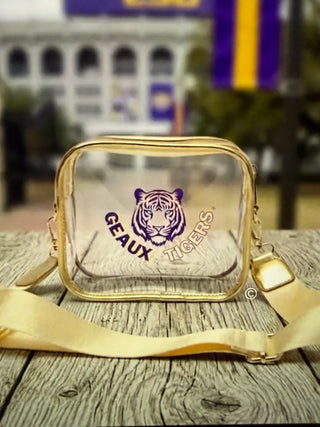 A clear rectangular crossbody bag with a gold metal frame and strap, featuring a purple LSU Geaux Tigers logo on the front, resting on a rustic wooden surface with a blurred outdoor background.