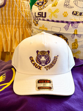 A white GAUX TIGERS baseball cap with a purple and gold embroidered tiger head logo and text, resting on a purple garment with LSU-themed bags in the background.