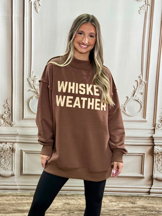 A woman wearing an oversized brown sweatshirt with the words 'WHISKE WEATHER' printed in beige across the chest, paired with black leggings, standing in front of an ornate cream-colored wall.