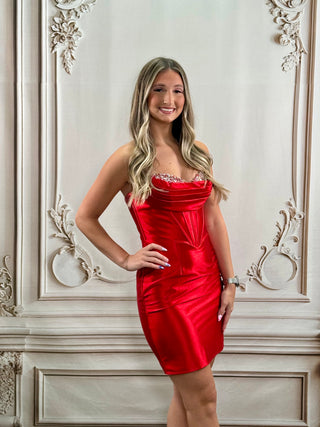 A woman modeling a bright red strapless satin mini dress with a sweetheart neckline and silver rhinestone trim, standing in front of an ornate beige wall.