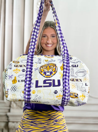 A woman holds up a large LSU Tigers themed duffel bag. The bag has a white and purple gingham pattern on the straps and sides, with a white front panel featuring the LSU tiger logo, 'LSU' text, and various LSU-themed graphics in purple, gold, and blue.