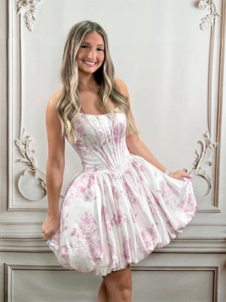 A woman modeling a sleeveless white dress with a pink floral print and a structured corset-style bodice, standing in front of an ornate light gray wall.