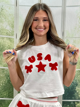 Woman wearing a white sleeveless shirt with red 'USA' and star designs, holding blonde hair.