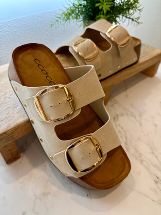 Beige sandals with gold buckles on a marble surface with a wooden bench and plant in the background.