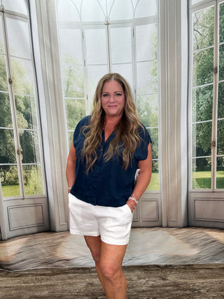 Woman in navy top and white shorts standing in a room with large windows.