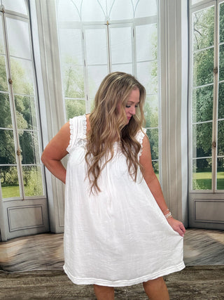 Woman wearing a white dress standing in a room with large windows.