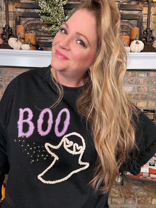 Woman wearing a black sweatshirt with 'BOO' and a ghost design, standing in front of a fireplace.