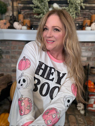 Woman wearing a 'Hey Boo' shirt with Halloween-themed designs in a decorated room.