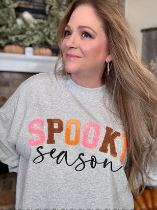 Woman wearing a gray sweatshirt with 'Spooky Season' text, standing in a room with decorative elements.