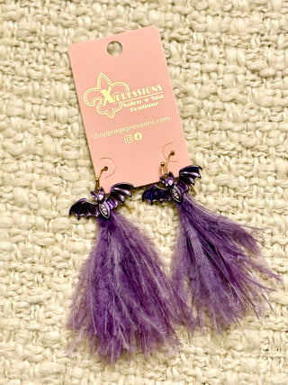 Purple feathered earrings with bat designs on a pink card against a textured beige background