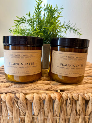 Two jars of 'Pumpkin Latte' candles from Jack Baker Candle Co. on a woven surface with a plant in the background.