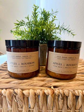 Two candles labeled 'Apples + Bourbon' by Jack Baker Candle Co. on a woven surface with a plant in the background.