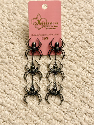 Pair of spider-shaped earrings on a pink card with 'Xpressions Salon & Spa Boutique' branding, displayed on a textured beige surface.