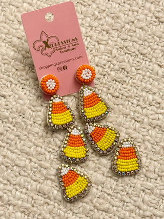 Candy corn-themed earrings on a pink card with 'Shopping Expressions' branding, on a textured beige surface.