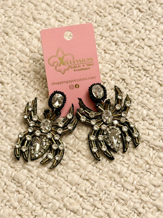 Pair of spider-shaped earrings with rhinestones on a textured beige surface, featuring a pink card with branding.