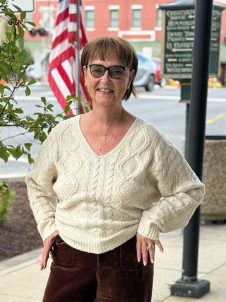 Woman wearing a cream sweater and glasses standing outdoors with an American flag in the background.