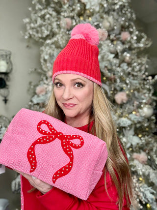 Woman wearing a pink knit hat and holding a pink gift box with a red bow in front of a decorated Christmas tree.