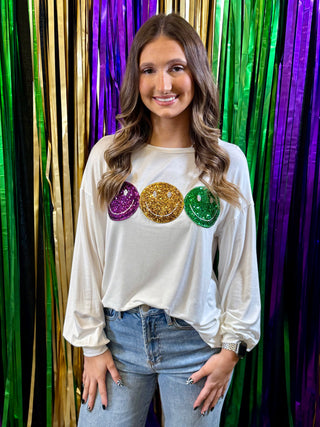 Woman wearing a white long-sleeve shirt with colorful sequin smileys in front of a colorful curtain.