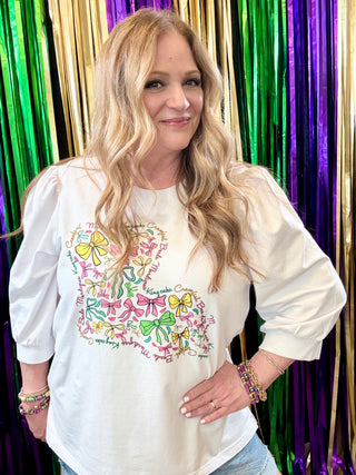 Woman wearing a white shirt with colorful designs in front of a colorful curtain.