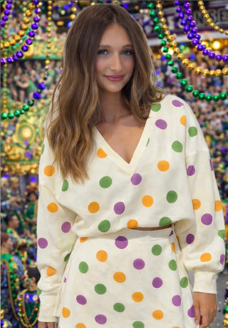 Woman standing in front of Mardi Gras beads wearing purple, green, and gold polka dot long sleeve, v-neck shirt with matching skirt