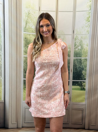 Woman wearing a pink floral dress standing in front of a glass door with a view of greenery.