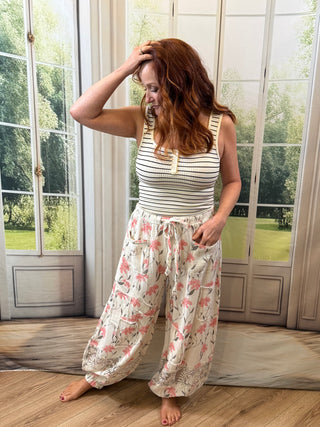 Woman wearing a striped tank top and floral pants standing in a room with large windows.