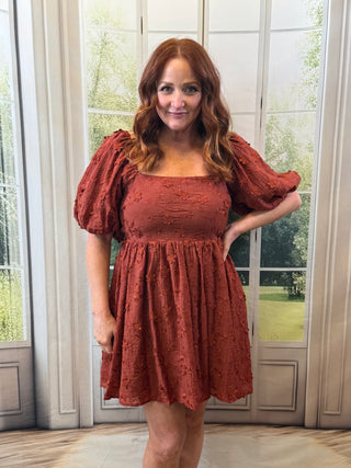 Woman wearing a rust-colored dress with puff sleeves in front of a window.